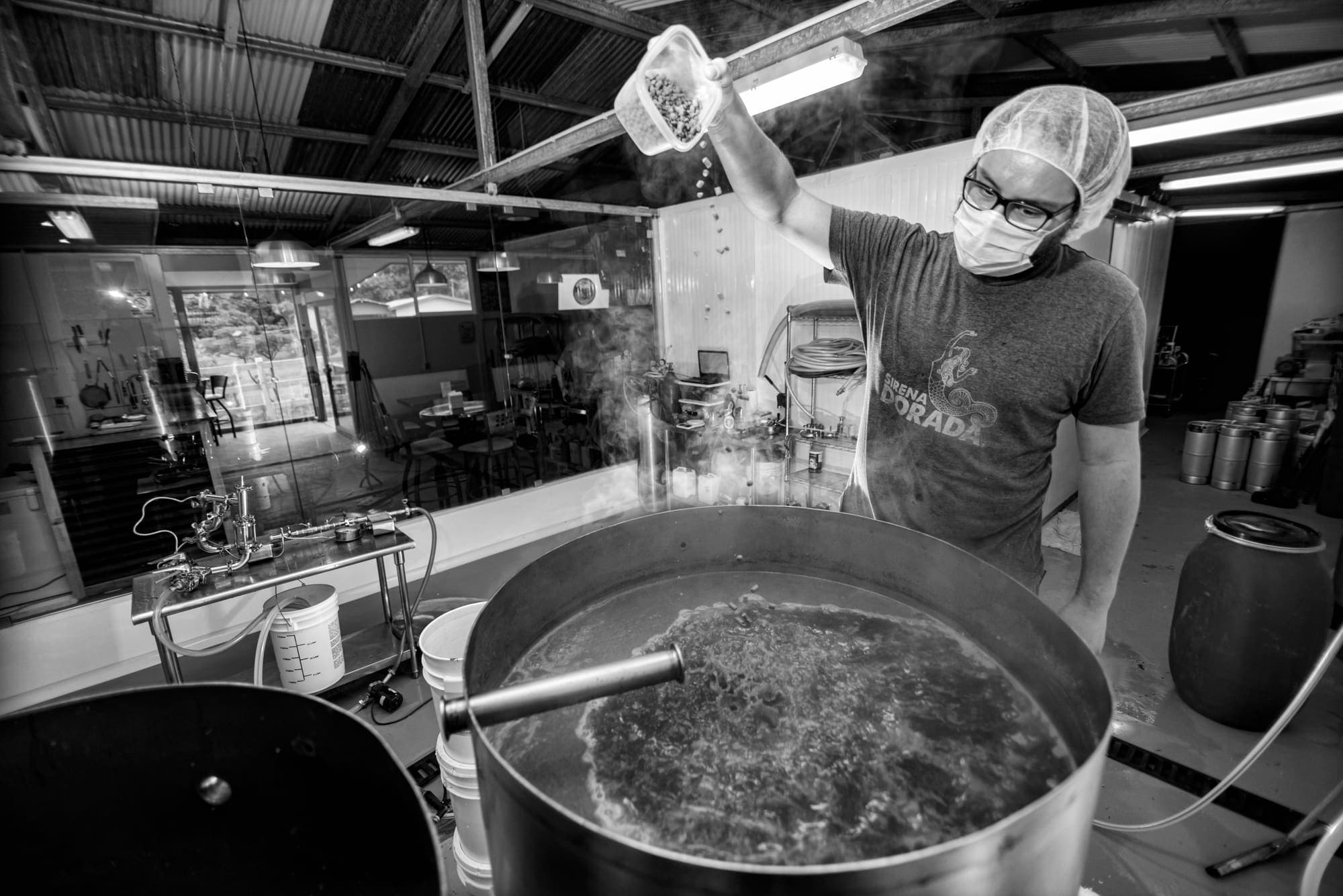 A brewer in a hairnet emptying a bag into the steaming kettle