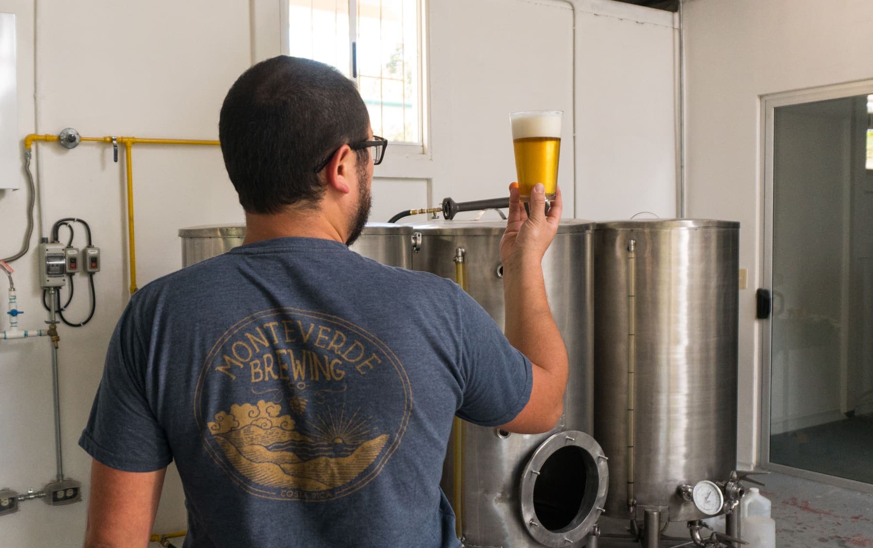 A brewer holds a glass of beer up to the light in front of the Monteverde Brewing Company brewhouse kettles